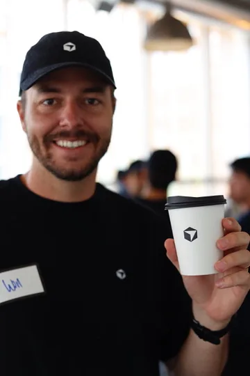 Will Rowston smiling at a Cursor event, wearing a Cursor cap and holding a branded coffee cup